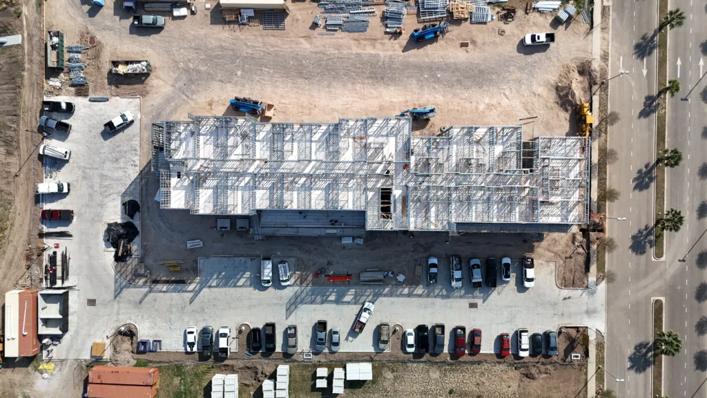 default - Scottsdale Construction Systems Aerial view of a construction site with a steel-framed building under construction, surrounded by equipment, material stacks, and a parking lot full of cars, next to a multi-lane road.