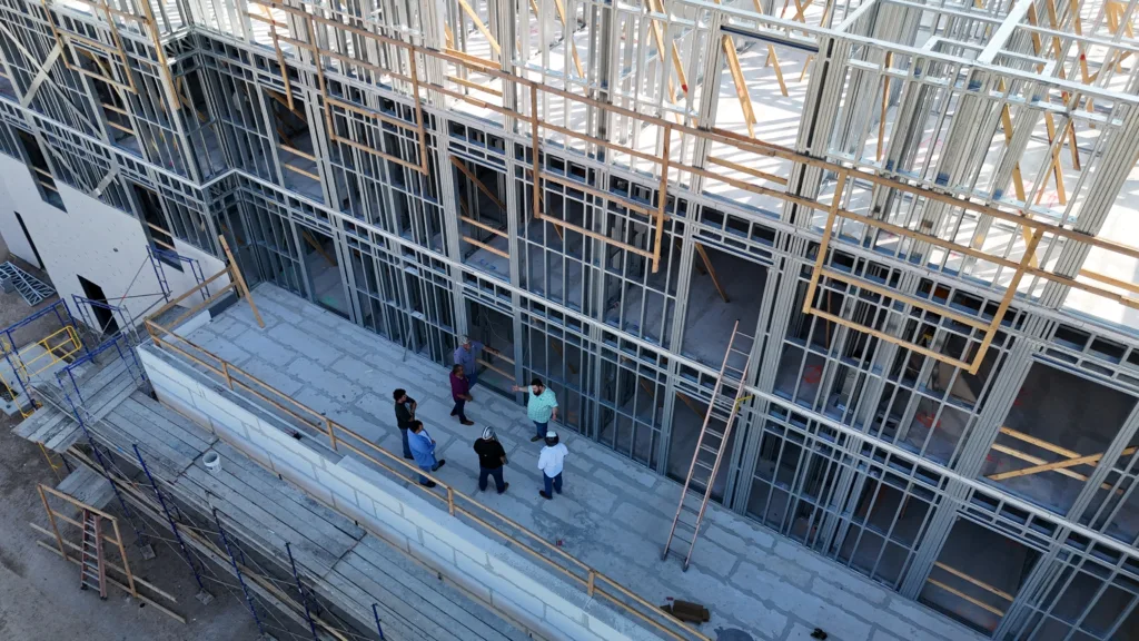 default - Scottsdale Construction Systems Under-construction building with exposed metal framing and wooden braces; a group of people stands on a concrete floor discussing around the skeletal structure