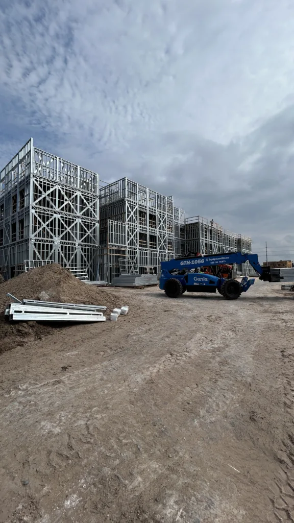 image00036 - Scottsdale Construction Systems Steel-framed building under construction on a dirt site, with a blue telehandler nearby and scattered metal pieces.