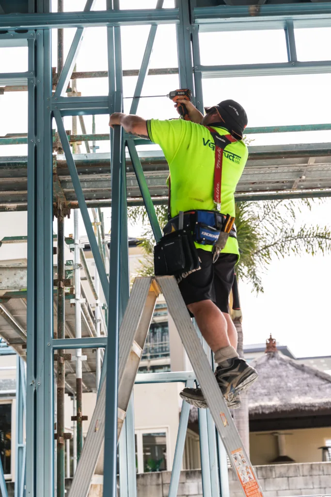 custom steel-framed coastal home 11 - Scottsdale Construction Systems Construction worker in a high-visibility shirt and helmet drills into blue steel scaffolding.