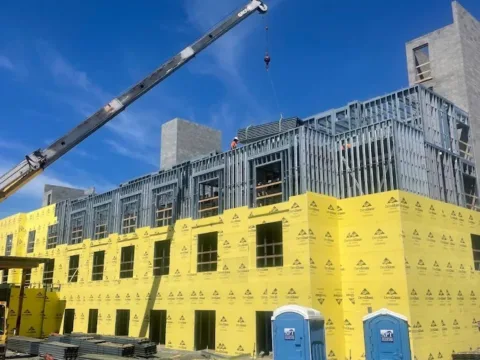 Construction site with a partially built yellow-wrapped building, a crane lowering a load, and blue portable toilets in the foreground.