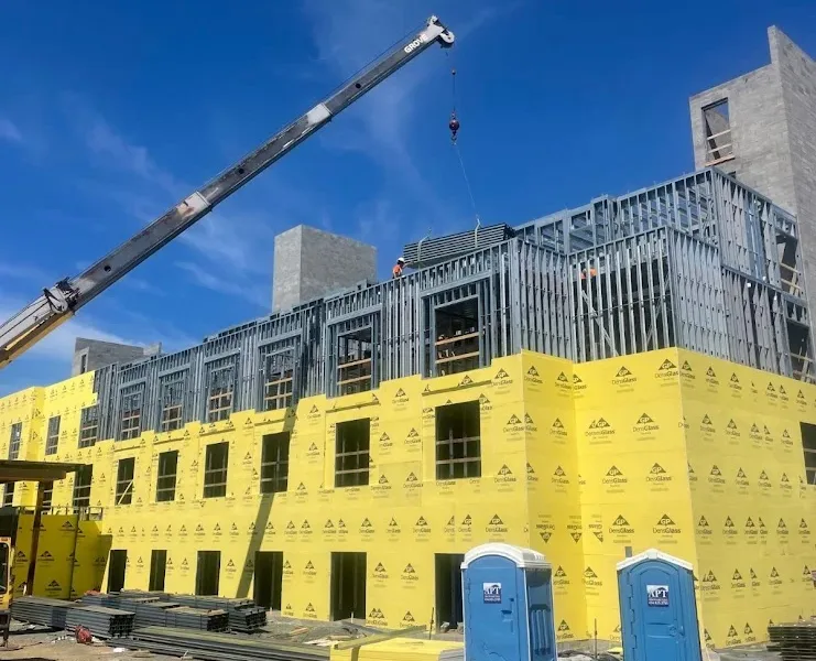 Construction site with a partially built yellow-wrapped building, a crane lowering a load, and blue portable toilets in the foreground.