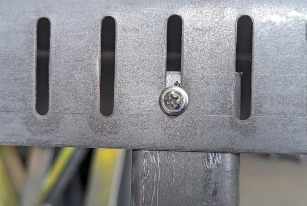 Low-profile pan-head screw riding in a vertical slot of galvanized slotted deflection track above a cold-formed steel stud, showing how slip track lets the floor above deflect, roll formed on a Scottsdale KFS Framemaker 1220G3 steel stud roll former, an Arkitech roll former alternative